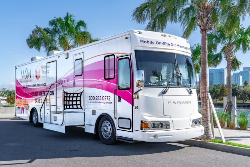 A mobile 3D mammography screening bus parked outdoors beside palm trees on a sunny day.