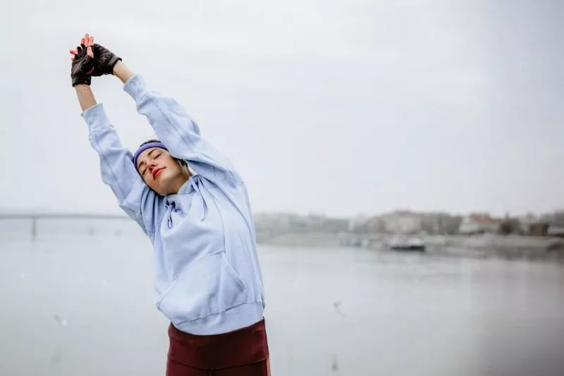 Woman stretching with arms raised by the water, wearing a hoodie and workout gloves on an overcast day.