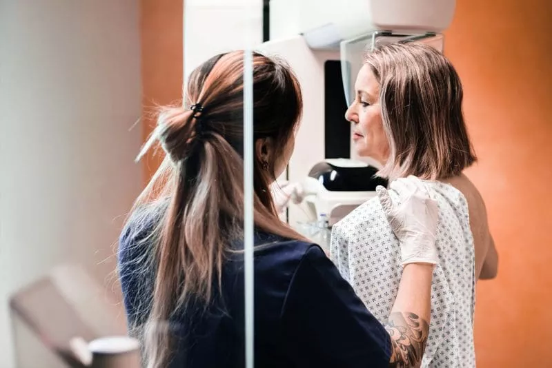 Patient standing at a mammography machine while a clinician gently positions her shoulder and upper body for the scan.