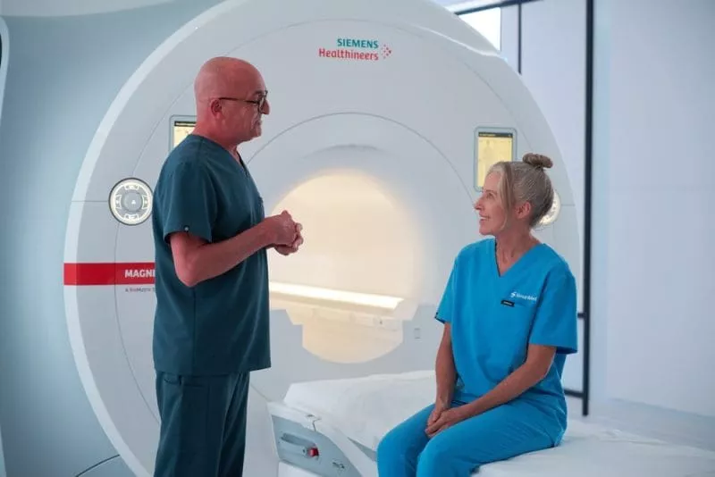 A patient sits on the edge of an MRI table while a clinician explains the procedure.