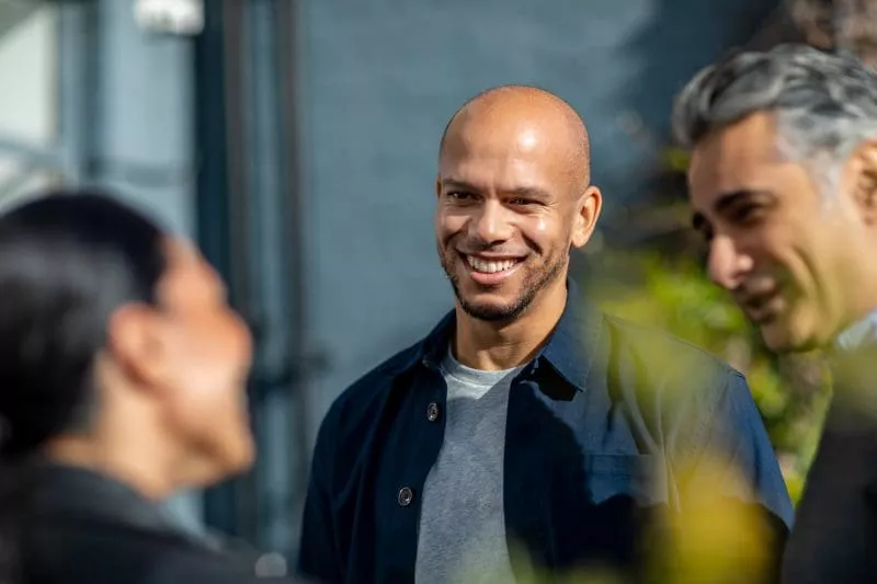Three people smiling and chatting outdoors, with one man in focus and two others blurred in the foreground.