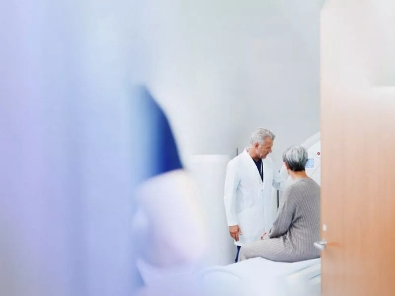 Doctor speaking with an older patient seated on an examination table in a clinical imaging room, viewed through a doorway.