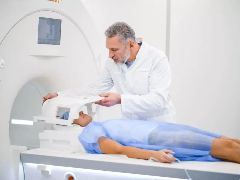 Clinician adjusting a head coil on a patient lying on an MRI scanner table in a bright clinical room.