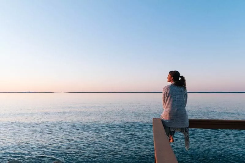 Person sitting on a wooden pier at sunset, looking out across calm water toward the horizon.