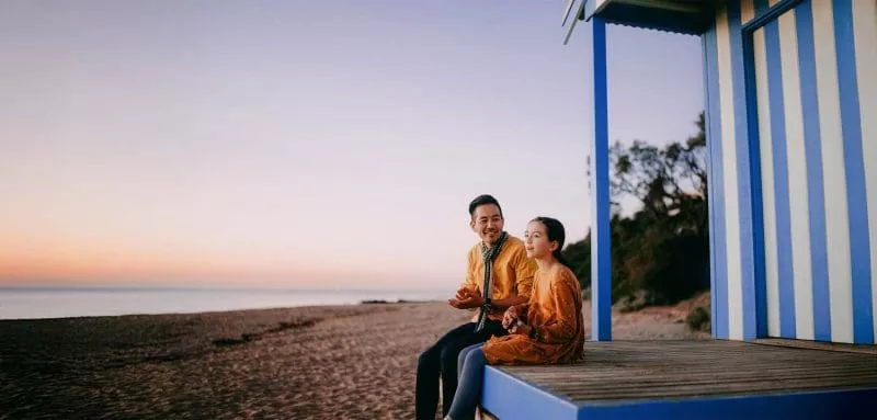 Adult and child sitting on a beach hut platform at sunset, talking and looking out towards the sea.