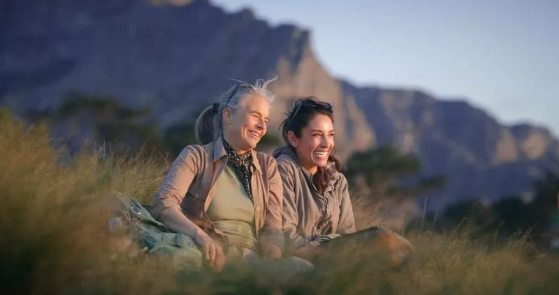 Two women sitting on grass outdoors, smiling together with mountains in the background at golden hour.