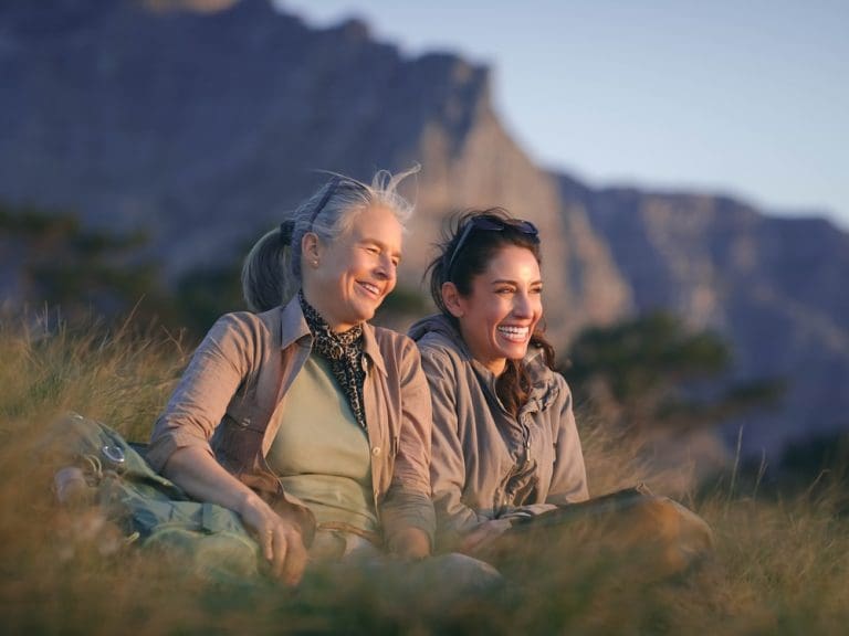 Two women sitting on grass outdoors, smiling together with mountains in the background at golden hour.