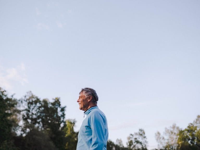 Older man in a blue jacket standing outdoors, looking to the side with trees and open sky behind him.