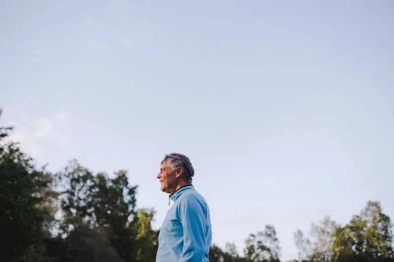 Older man in a blue jacket standing outdoors, looking to the side with trees and open sky behind him.
