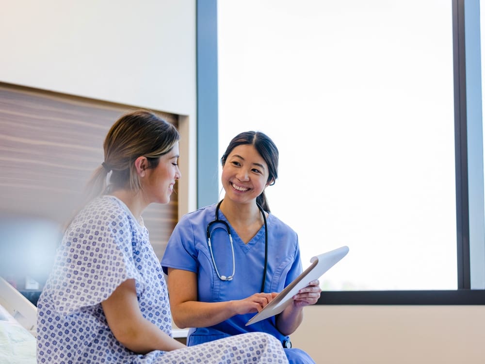 Nurse in blue scrubs smiling while reviewing a tablet with a patient sitting on a hospital bed.