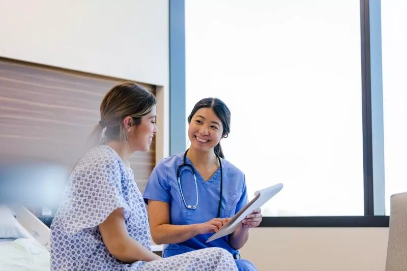 Nurse in blue scrubs smiling while reviewing a tablet with a patient sitting on a hospital bed.