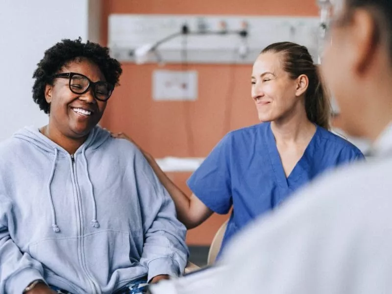 Smiling patient in a hoodie sitting with two healthcare professionals, one gently resting a hand on her shoulder in a hospital room.