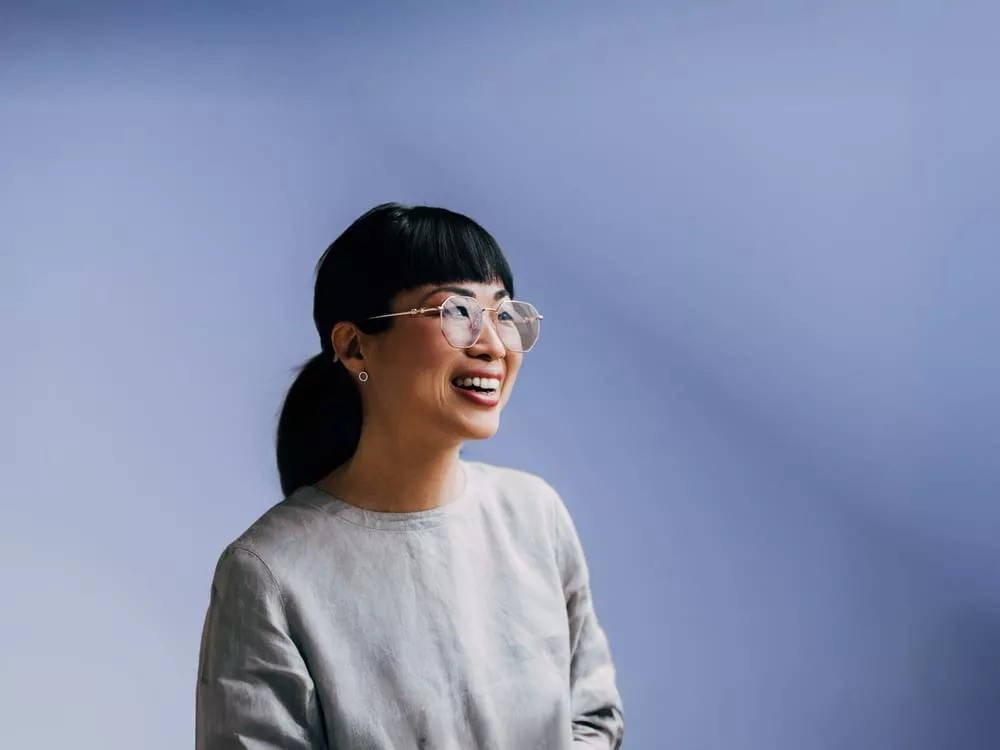 Smiling woman with glasses and a low ponytail looking to the side against a soft blue background.