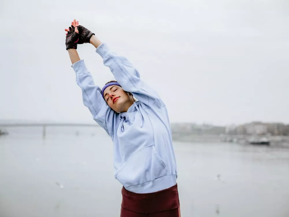 Person stretching with arms raised overhead by the water, wearing a hoodie and gloves.