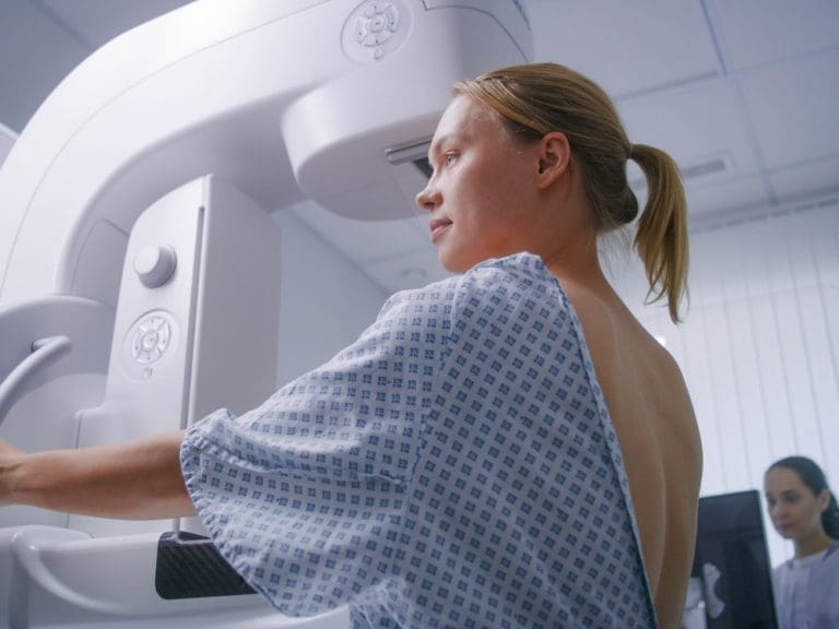 Patient positioning herself at a mammography machine while a technician stands nearby in the background.