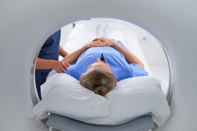 Patient lying on an MRI scanner bed, viewed from inside the scanner opening, with a clinician standing beside them.
