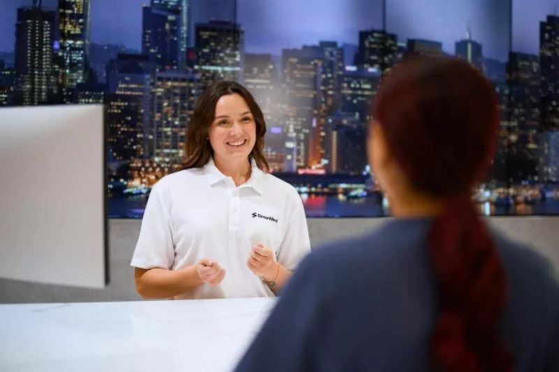 Clinic receptionist smiling and speaking with a clinician at the front desk, with a city skyline display in the background.