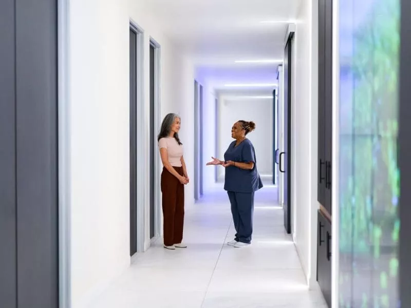 A clinician speaks with a woman in a bright, modern clinic corridor lined with consultation rooms.