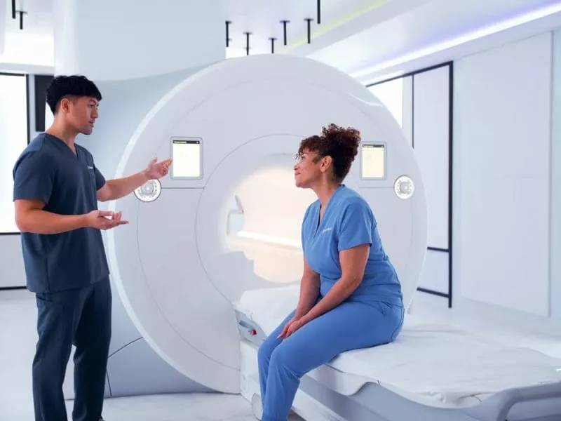 A clinician explains an MRI scan to a patient seated on the scan table beside the machine.