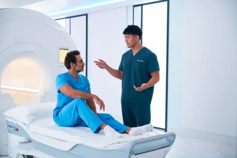 Two clinicians talking beside an MRI scanner, one seated on the scan table in a modern imaging room.